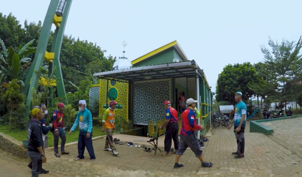 Group of cyclists in colorful jerseys chat outside a green-and-yellow pavilion with decorative lattice walls in a park.