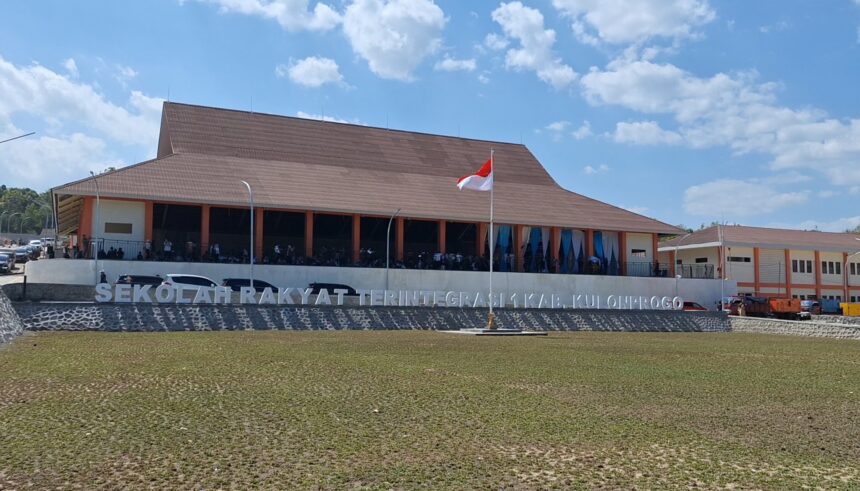 Front view of a large brown-roofed building with an Indonesian flag, people on the shaded veranda, and a stone wall with large letters in front.