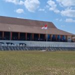 Large school building with a brown sloped roof and orange pillars, Indonesian flag raised on a pole, people gathered on the front veranda.