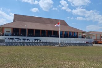 Large school building with a brown sloped roof and orange pillars, Indonesian flag raised on a pole, people gathered on the front veranda.