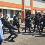 Group of uniformed trainees performing a martial arts drill outdoors, supervised by a man in a white coat and black hat, with onlookers in the background.