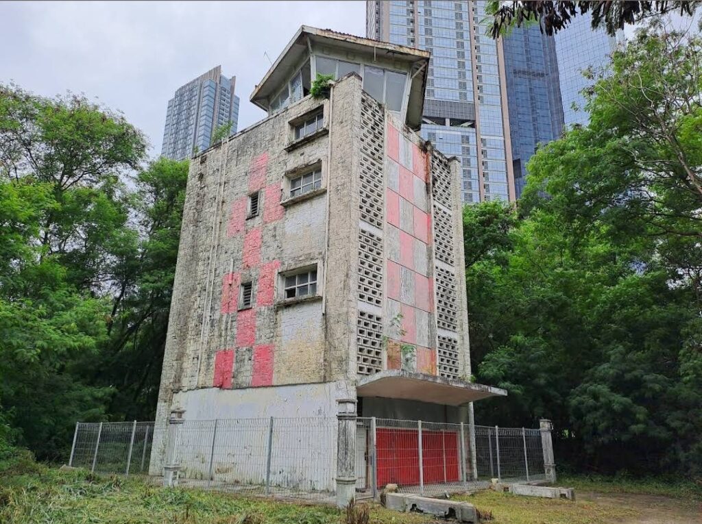 Dilapidated concrete building with red panels, fenced yard, set in a green park with modern skyscrapers in the background.