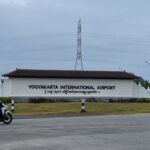 Yogyakarta International Airport sign with a statue to the left and a motorcyclist passing on the road in the foreground.