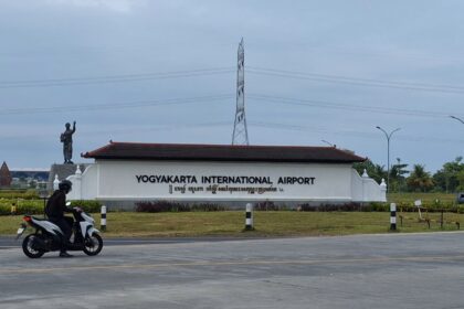 Yogyakarta International Airport sign with a statue to the left and a motorcyclist passing on the road in the foreground.