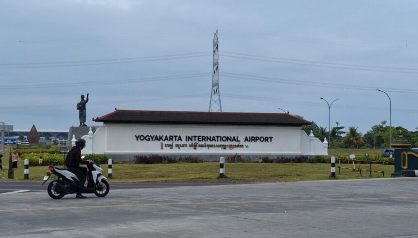 Yogyakarta International Airport sign with a statue to the left and a motorcyclist passing on the road in the foreground.