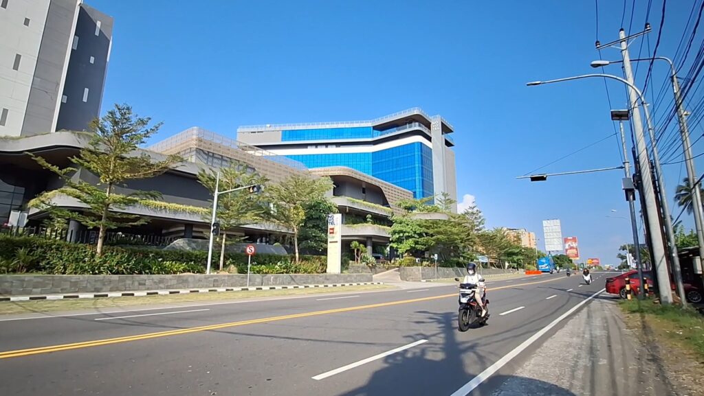Wide street with a modern blue-glass office building on the left under a clear blue sky; foreground shows a motorcyclist riding toward the camera near a landscaped median. A row of utility poles and power lines runs along the right side of the scene.