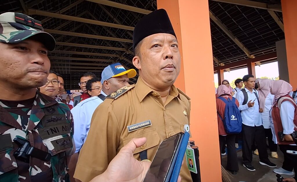 Indonesian official in brown uniform being interviewed by reporters, with a camouflaged soldier on the left and students in the background under a covered area.