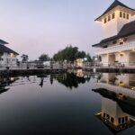 White, multi-story buildings with dark roofs line a walkway by a calm canal at dusk, their reflections visible in the water.