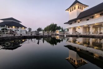 White, multi-story buildings with dark roofs line a walkway by a calm canal at dusk, their reflections visible in the water.