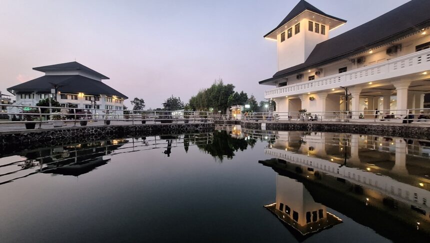 White, multi-story buildings with dark roofs line a walkway by a calm canal at dusk, their reflections visible in the water.