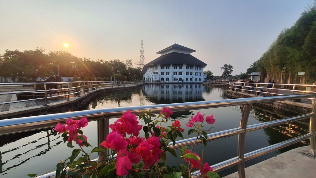 Sunset over a pond with a white multi-story building in the center, pink flowers in the foreground, and people walking along the railing.