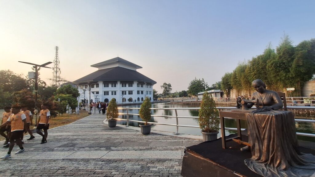 Bronze statue of a seated monk playing a drum on a stage beside a railing and pond, with a white multi-story building in the background.