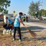 Three workers in blue uniforms sweep gravel along a roadside market area with water jugs and bags nearby