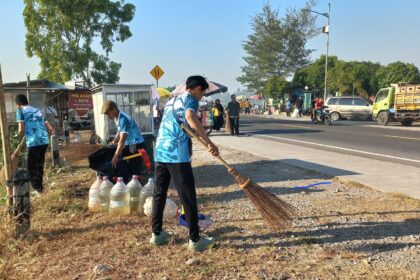 Three workers in blue uniforms sweep gravel along a roadside market area with water jugs and bags nearby