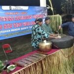 Group of men in traditional clothing performing a ritual with large cauldron and wooden tools on a decorated stage, banner in Indonesian in the background