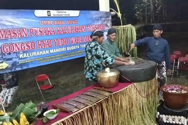 Group of men in traditional clothing performing a ritual with large cauldron and wooden tools on a decorated stage, banner in Indonesian in the background