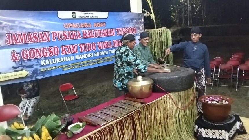 Group of men in traditional clothing performing a ritual with large cauldron and wooden tools on a decorated stage, banner in Indonesian in the background