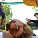 Group of people in traditional clothing performing a ceremonial drum beat on a large wooden drum.