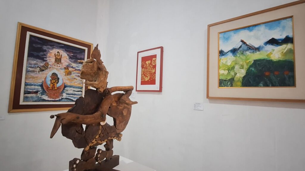 Wooden abstract sculpture centered in an art gallery with framed paintings on the wall behind it.