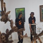 Two men in black shirts with orange logos stand beside large driftwood sculptures and a ship model in an art gallery.