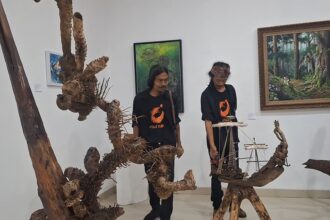 Two men in black shirts with orange logos stand beside large driftwood sculptures and a ship model in an art gallery.