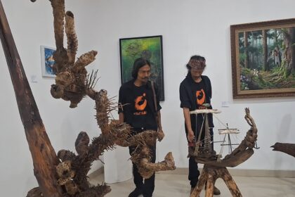 Two men in black shirts with orange logos stand beside large driftwood sculptures and a ship model in an art gallery.