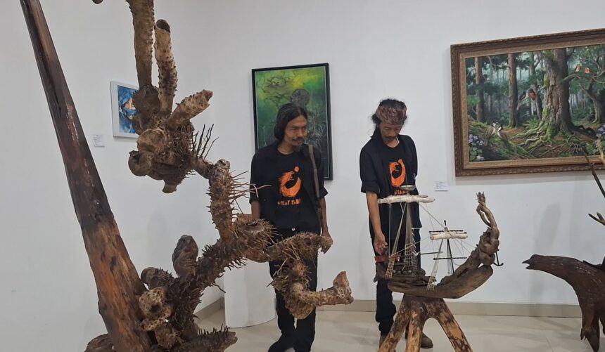Two men in black shirts with orange logos stand beside large driftwood sculptures and a ship model in an art gallery.