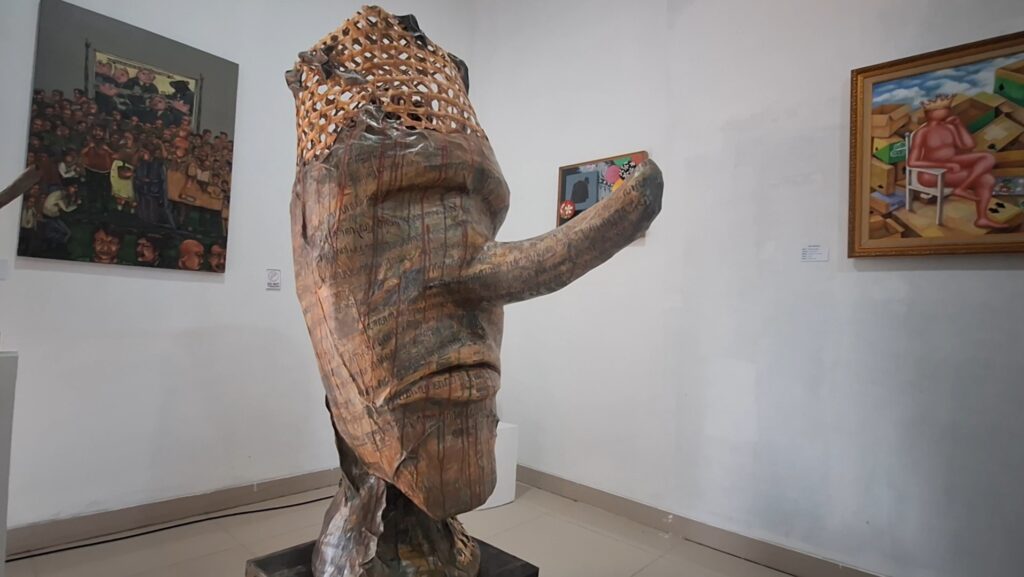 Large wooden sculpture of a twisting human figure with a lattice head in a gallery, outstretched arm pointing forward, surrounded by framed paintings on white walls.