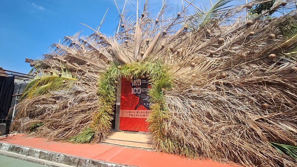 Entrance framed by a large dried palm frond installation with a red poster behind the doorway.