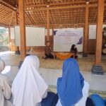Man with microphone speaks at a community meeting in a wooden hall, with a woman beside a banner at the front and attendees in hijabs watching.