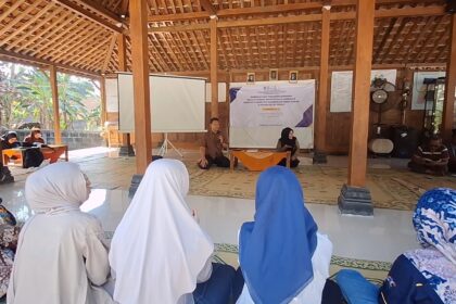Man with microphone speaks at a community meeting in a wooden hall, with a woman beside a banner at the front and attendees in hijabs watching.