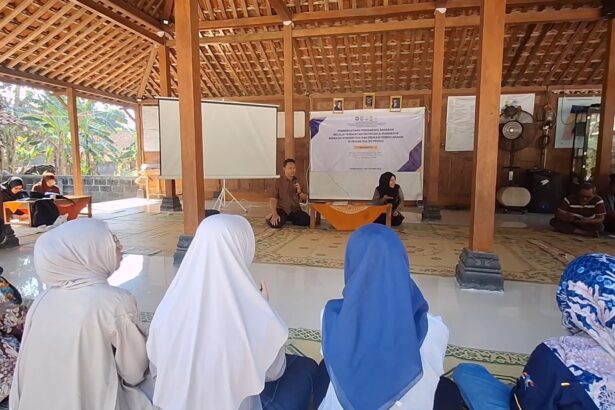 Man with microphone speaks at a community meeting in a wooden hall, with a woman beside a banner at the front and attendees in hijabs watching.