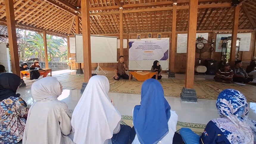 Screenshot_20260713_091334_Gallery - Mabur.co Man with microphone speaks at a community meeting in a wooden hall, with a woman beside a banner at the front and attendees in hijabs watching.