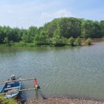 Blue wooden boat with oars moored at the rocky riverbank, calm water and dense green trees beyond under a blue sky.