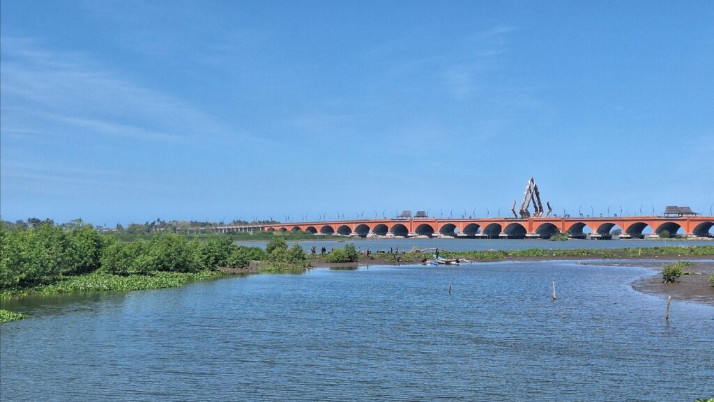 Screenshot_20260713_183404_Gallery - Mabur.co Orange arched bridge spanning a calm river beneath a clear blue sky, with a triangular sculpture at the center and people near the shore.