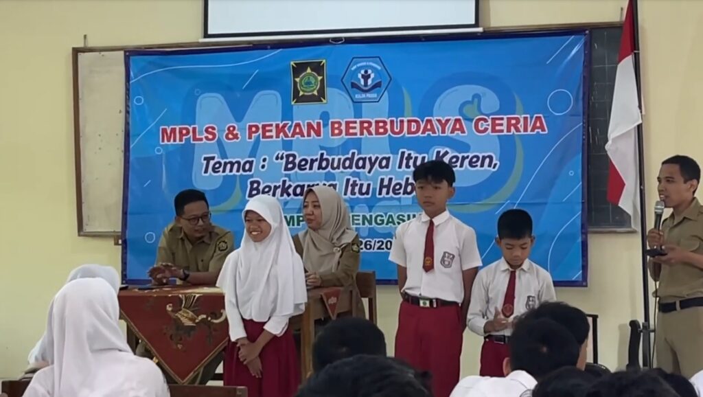 School event on stage with students in white shirts and red skirts, a blue banner behind them reading MPLS & Pekan Berbudaya Ceria, and the Indonesian flag on the right.
