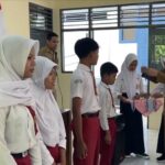 Teacher in a tan uniform hands a ribboned gift to a student during a school ceremony, with classmates lined up in white hijabs and red uniforms nearby.