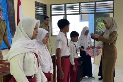 Teacher in a tan uniform hands a ribboned gift to a student during a school ceremony, with classmates lined up in white hijabs and red uniforms nearby.