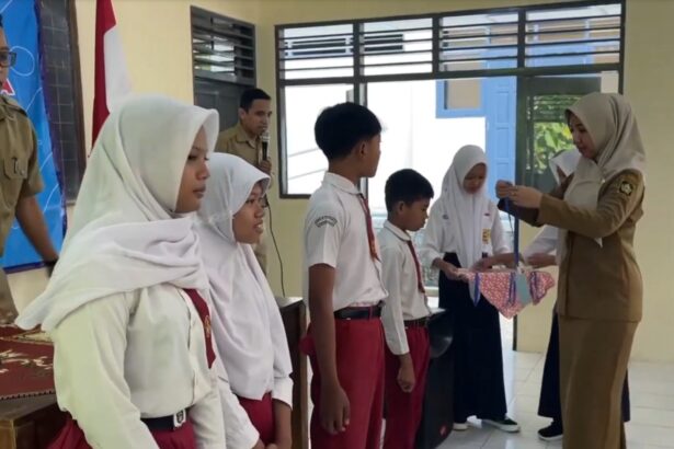 Teacher in a tan uniform hands a ribboned gift to a student during a school ceremony, with classmates lined up in white hijabs and red uniforms nearby.