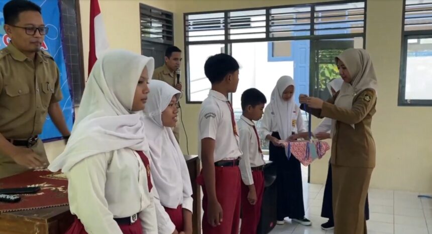 Teacher in a tan uniform hands a ribboned gift to a student during a school ceremony, with classmates lined up in white hijabs and red uniforms nearby.