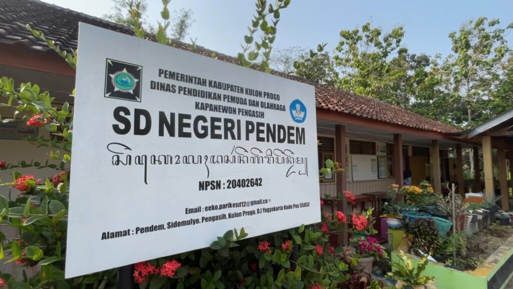 Screenshot_20260715_084302_Gallery - Mabur.co Large outdoor signboard for SD Negeri Pendem school with Indonesian text and logos, surrounded by plants and a building in the background.