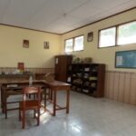 An empty classroom with wooden desks and chairs, shelves along the back wall, and windows letting in light.