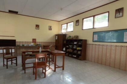 An empty classroom with wooden desks and chairs, shelves along the back wall, and windows letting in light.