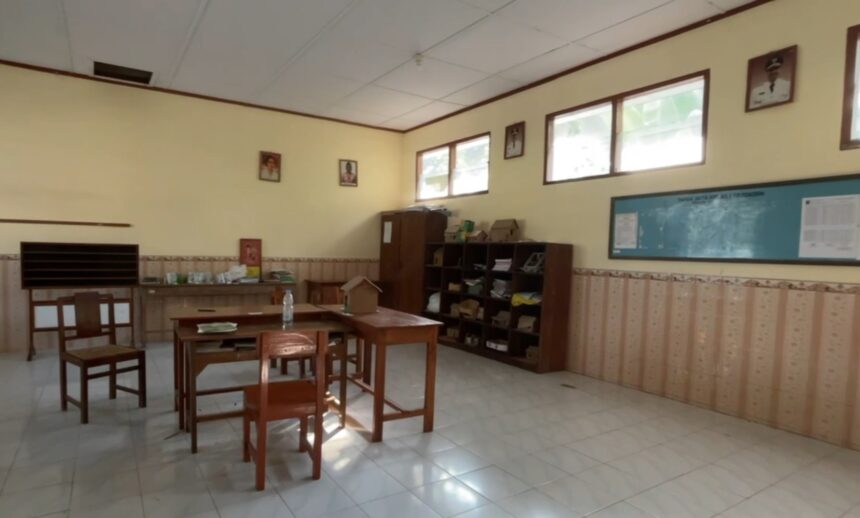 An empty classroom with wooden desks and chairs, shelves along the back wall, and windows letting in light.