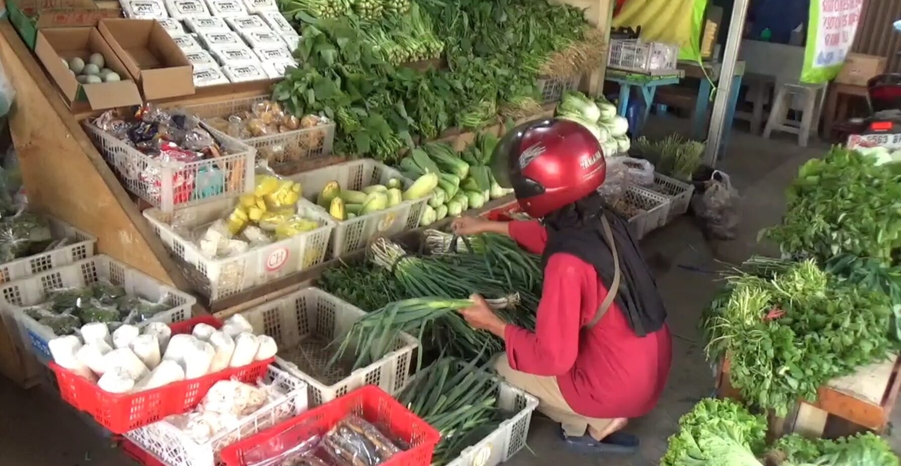 Person wearing a red shirt and helmet selecting long green onions from a market stand with crates of vegetables and herbs around.