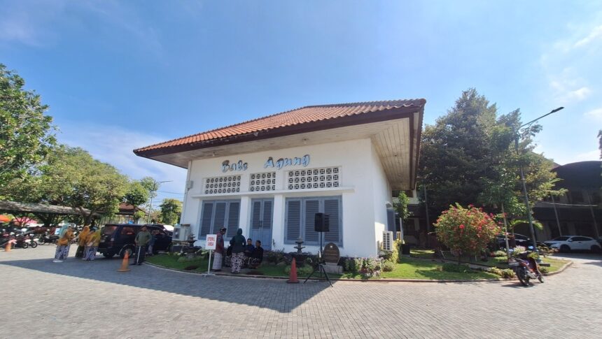 White building with a red-tiled roof and blue shutters, people gathered outside in a paved courtyard with trees nearby.