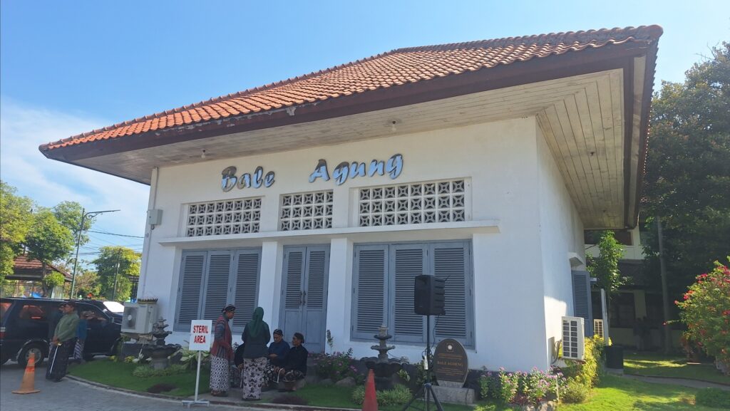 Screenshot_20260715_095128_Gallery - Mabur.co Front view of a white Balinese community hall labeled 'Bale Agung' with a red tile roof; people stand and sit outside near a 'Steril Area' sign on a sunny day.