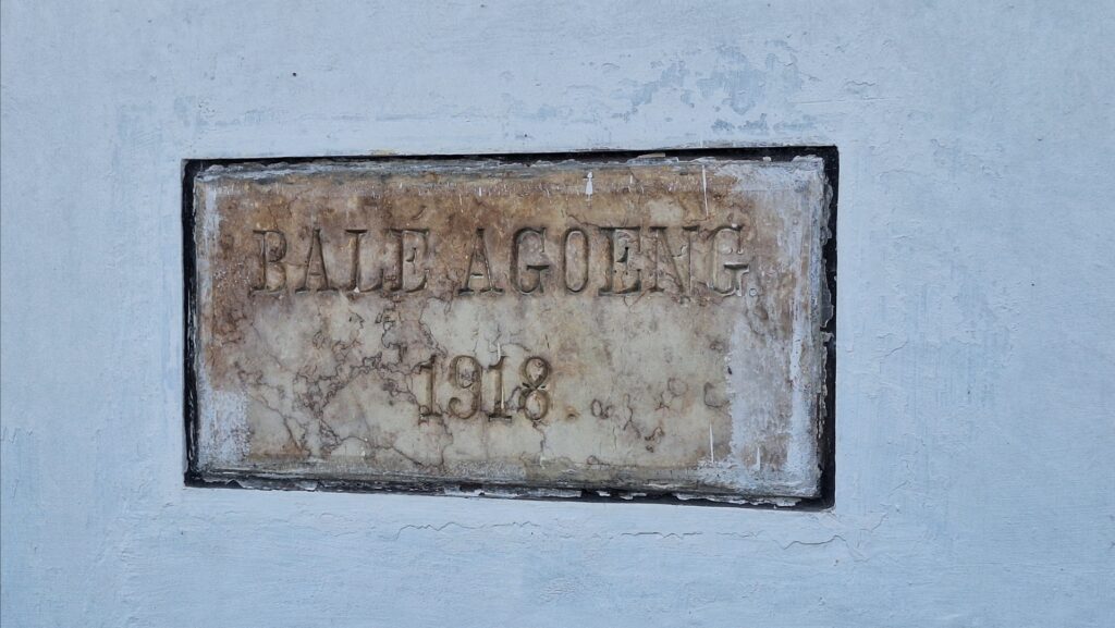 Screenshot_20260715_095143_Gallery - Mabur.co Weathered rectangular plaque embedded in a blue wall, with raised letters that appear to read 'BALE AGENG' and the year '1918' in the lower portion.