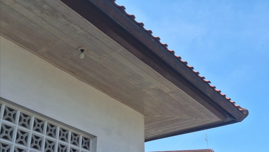 Screenshot_20260715_095208_Gallery - Mabur.co Corner of a building with a tiled sloped roof, exposed light bulb, and decorative block wall against a clear blue sky.