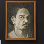 Framed charcoal portrait of a man with short hair and mustache, on a dark gallery wall.